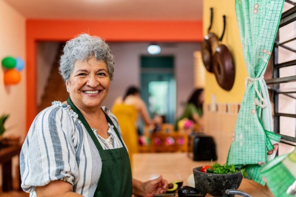 An older adult woman wearing an apron is preparing food in her kitchen and smiling at the camera.