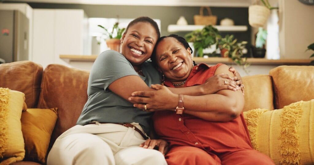 A woman and her older adult mother sit on a couch, hugging each other and smiling at the camera.