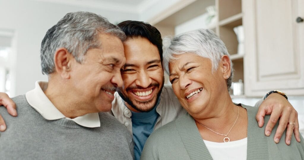 A man stands behind his older adult mother and father with his arms around their shoulders. They are all smiling.