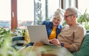 A smiling older adult woman and man sit on a couch together looking at a laptop.