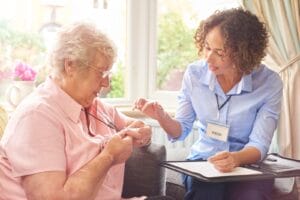 A woman shows an older adult woman how to use her medical alert system button, which the older woman is wearing around her neck.