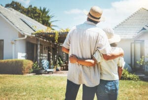 An image of a couple standing arm in arm on a sunny day, standing with their backs to the camera. They look at the backyard and home in front of them.