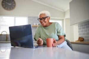An older adult woman sits in her kitchen with a laptop and a coffee mug on the counter in front of her.