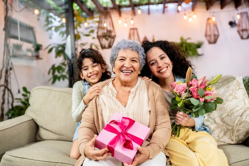 An older adult woman is holding a wrapped present. She is sitting on a couch with her daughter and granddaughter. The daughter is holding a bouquet of flowers.