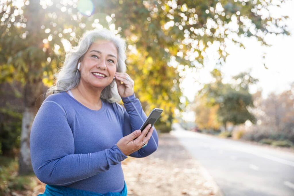 A woman is outside walking. She is putting an earbud in her ear and holding a smartphone.