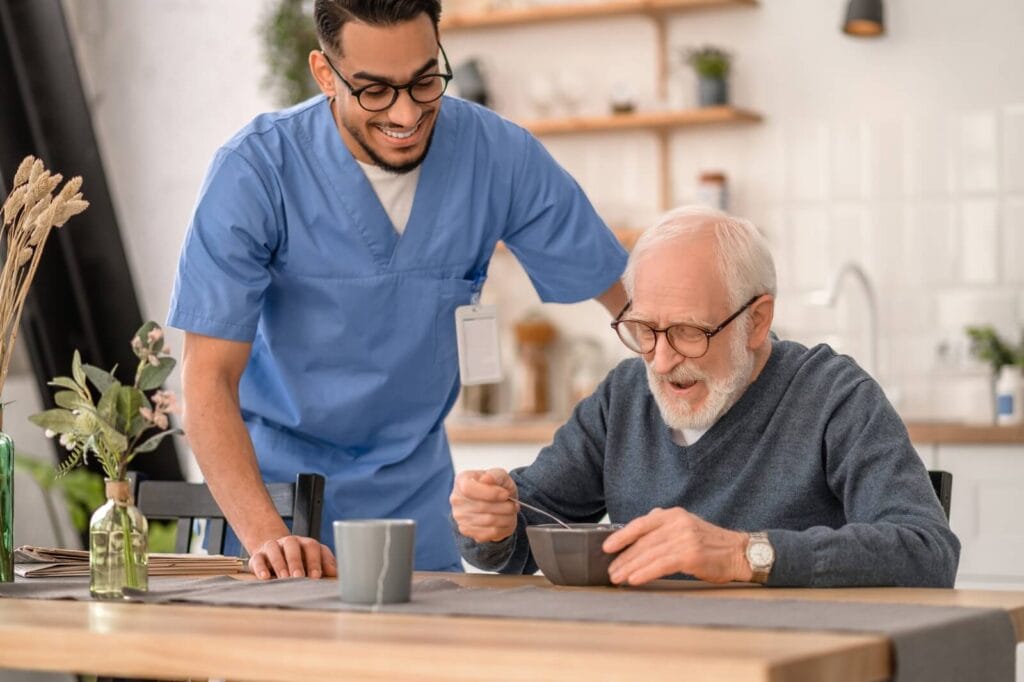 An older adult man sits at a kitchen table, eating from a bowl. A male caregiver stands beside him.