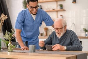 An older adult man sits at a kitchen table, eating from a bowl. A male caregiver stands beside him.