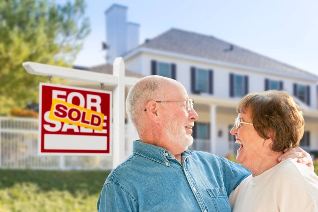 An older adult man and woman are standing in front of a house and a for sale sign that says "sold." They are smiling at each other.
