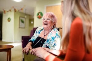 An older adult woman wearing a nasal cannula sits next to a younger woman. She is laughing.