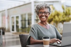 An older adult woman sits at a table with a laptop and a disposable coffee cup in front of her. She smiles at the camera.