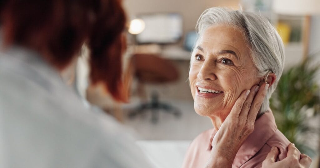 An older adult woman wearing a hearing aid is speaking to someone shown from behind.