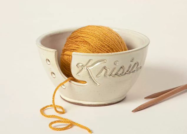 Product image of a tan ceramic yarn bowl personalized with a person's name. The bowl contains a ball of yellow yarn to demonstrate how to use it and the bottom of two knitting needles.