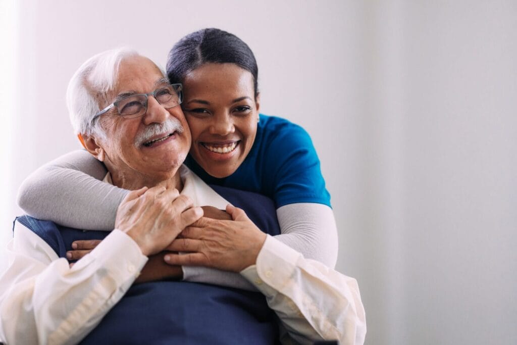 An older adult sits while a female caregiver hugs him from behind the chair. They smile.