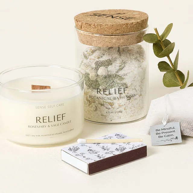 Photo of a jar of bath soak, which includes salt and botanicals, a rosemary-and-sage-scented candle, a tea bag, and a box of matches. There is also a sprig of eucalyptus in the photo.