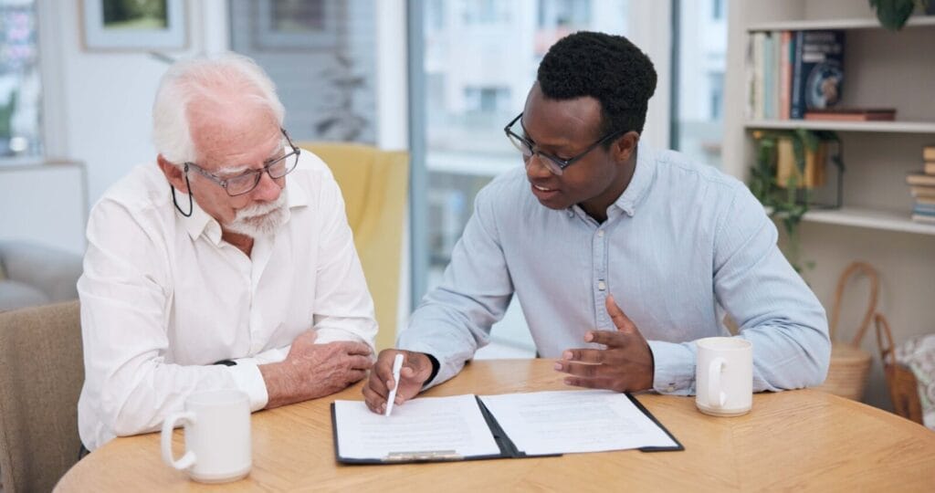 An older adult man sits at a table with a younger man, looking over paperwork in a folder.