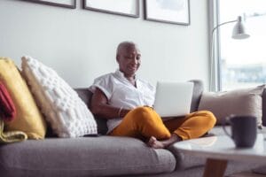 An older adult woman sits on a couch with her legs tucked under her. She has a laptop on her lap and is smiling.