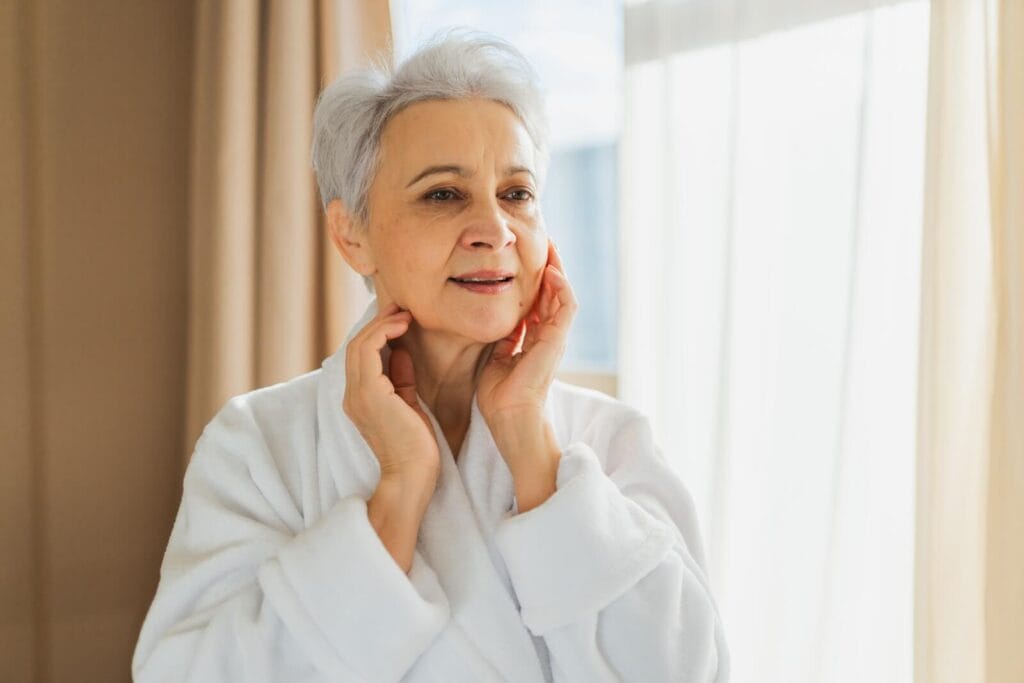 An older adult woman wearing a bathrobe has her hands up to her face.