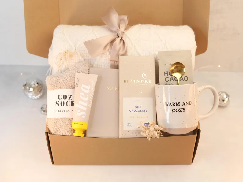 Photo of a gift box that holds cozy socks, shea butter moisturizer, a notebook, a bar of milk chocolate, a mug that reads "WARM AND COZY," a brass-colored spoon, a package that reads "HOT CACAO," and a woven blanket.