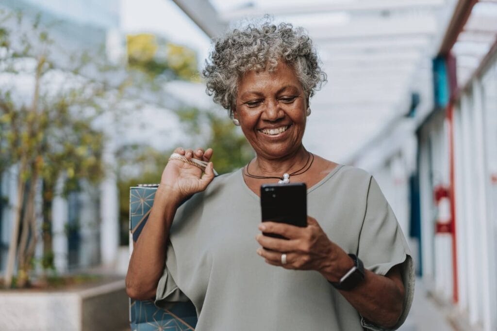 An older adult woman is standing and looking at her cell phone. She is smiling.