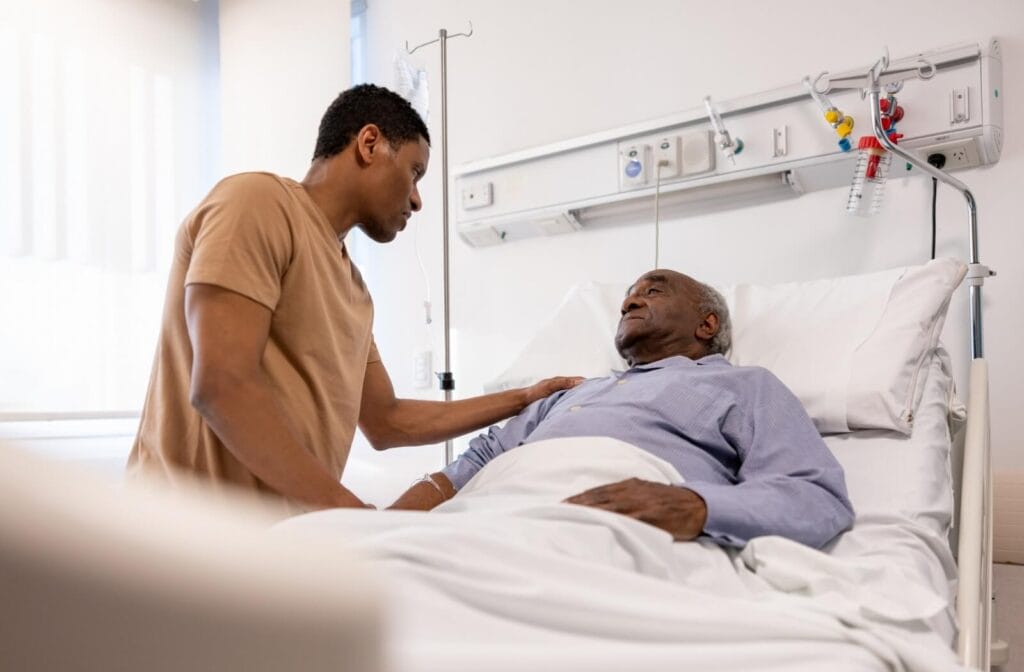 A man stands beside his older adult father's hospital bed. He has his hand on his shoulder.