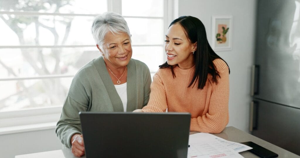An older adult woman and her adult daughter sit at a table with a laptop computer and paperwork on the table in front of them.