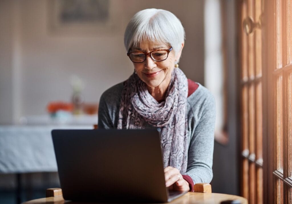 An older adult woman sits at a table with a laptop in front of her.