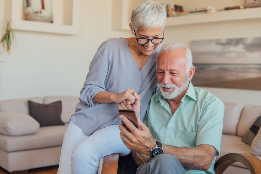 An older adult woman and man sit huddled together, looking at a smartphone that the man is holding.