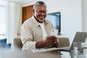 An older adult man is smiling while looking at his smartphone. There is also a laptop on the table in front of him.