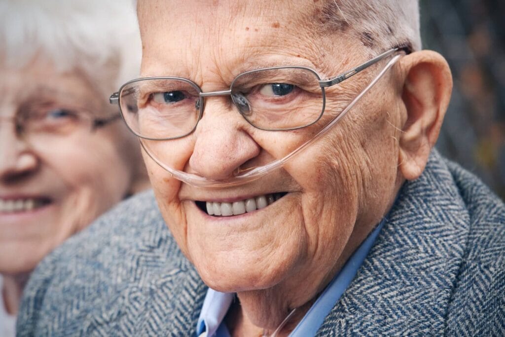 An older adult man wearing a nasal cannula smiles at the camera. An older adult woman is in the background,