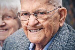 An older adult man wearing a nasal cannula smiles at the camera. An older adult woman is in the background,