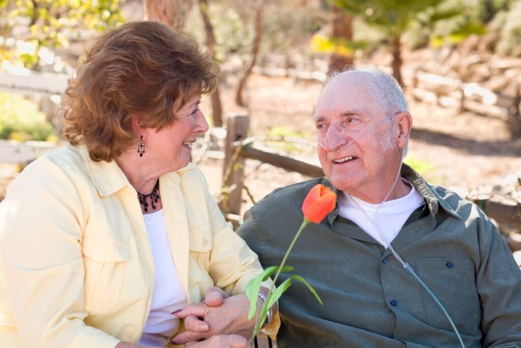 An older adult woman and man are sitting outdoors. He is wearing a nasal cannula for supplemental oxygen. She is holding a tulip.