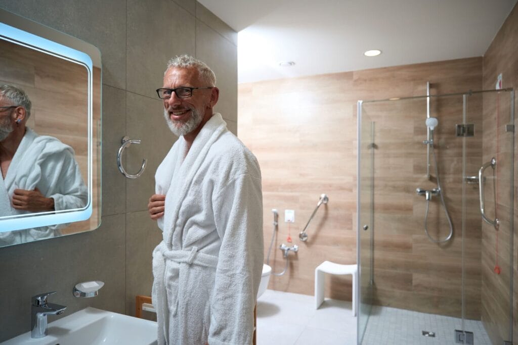 An older adult man wearing a bathrobe stands in a bathroom. There is a walk-in shower behind him. He smiles at the camera.