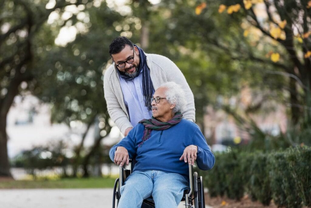 A man is pushing his older adult father in a wheelchair. They are outside.