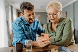A man sits at a counter with his older adult mother, showing her a cell phone.