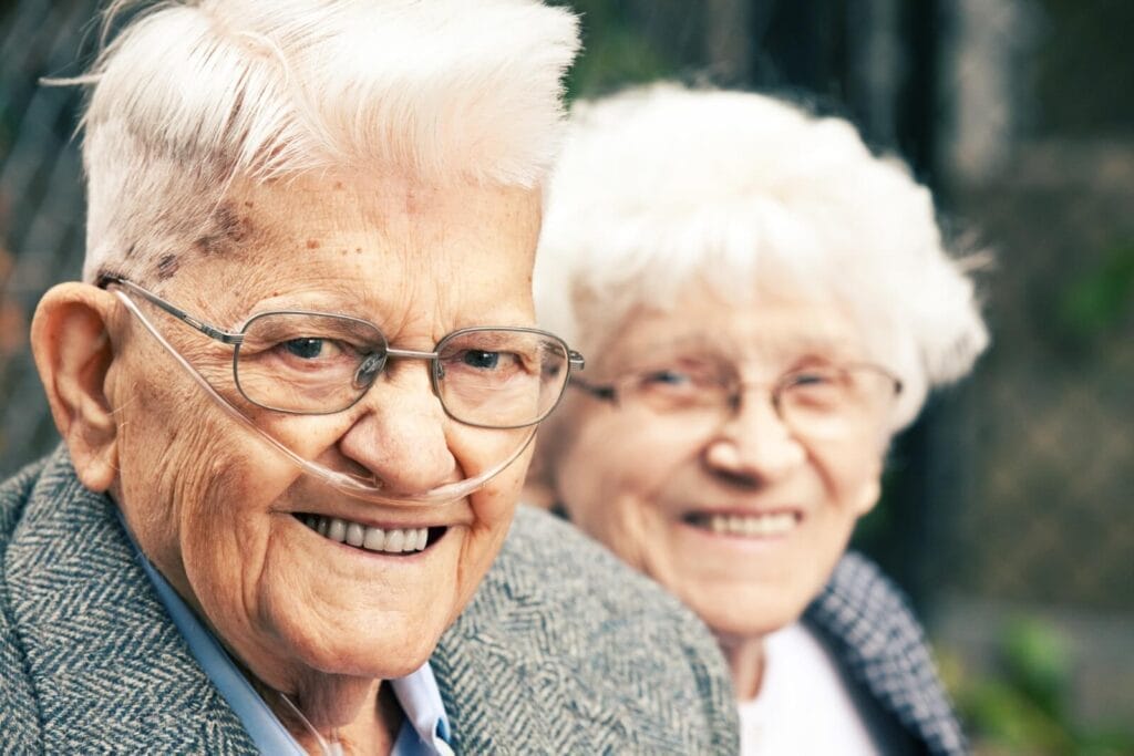 Close-up of an older adult man and woman smiling at the camera. The man has a nasal cannula for oxygen.