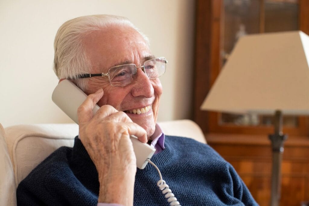 An older adult man is talking on a landline telephone with a cord.