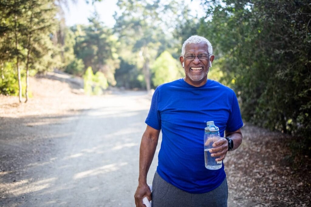 An older adult man is outside on a walking path. He is holding a bottle of water and smiling at the camera.