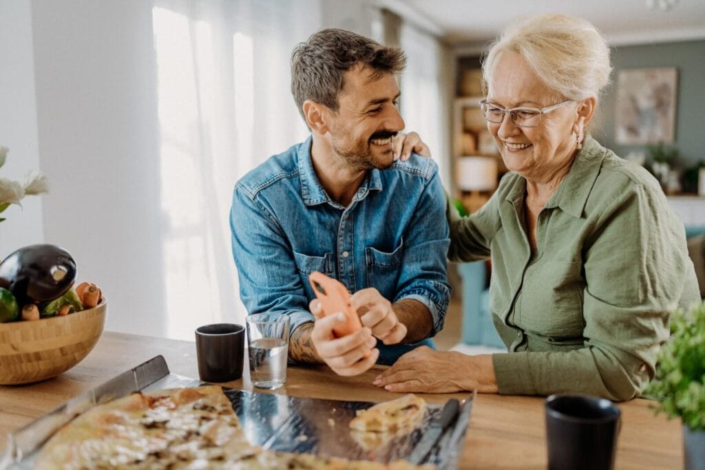 A man is standing at a counter with his older adult mother, showing her a smartphone. They are smiling, and she has her hand on his shoulder. There is a pizza and some glasses in front of them.