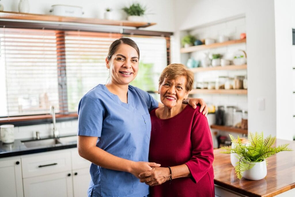 A female caregiver wearing scrubs and an older adult woman are standing in a kitchen with their arms around each other. They smile at the camera.