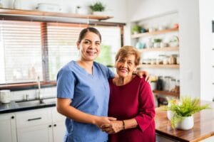 A female caregiver wearing scrubs and an older adult woman are standing in a kitchen with their arms around each other. They smile at the camera.