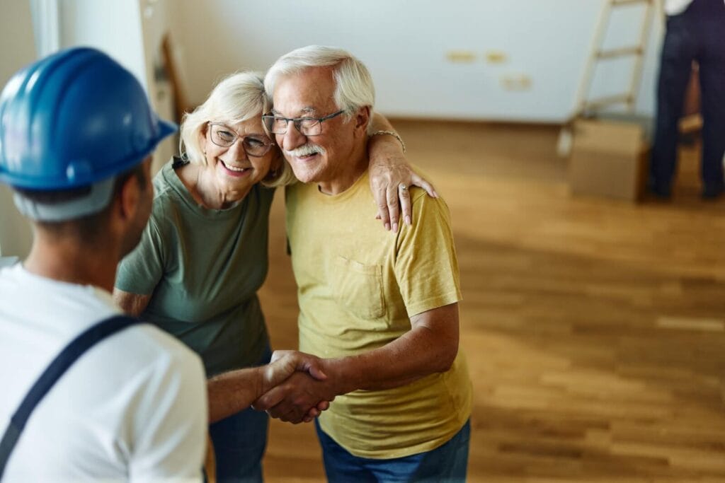 A man shown from behind is wearing a hard hat and shaking the hand of an older adult man, who has his other arm around an older adult woman. They are smiling.