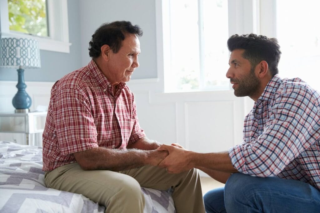 A man is crouching in front of his older adult father, who is sitting on a bed. He is holding both of his father's hands. They are looking at each other.