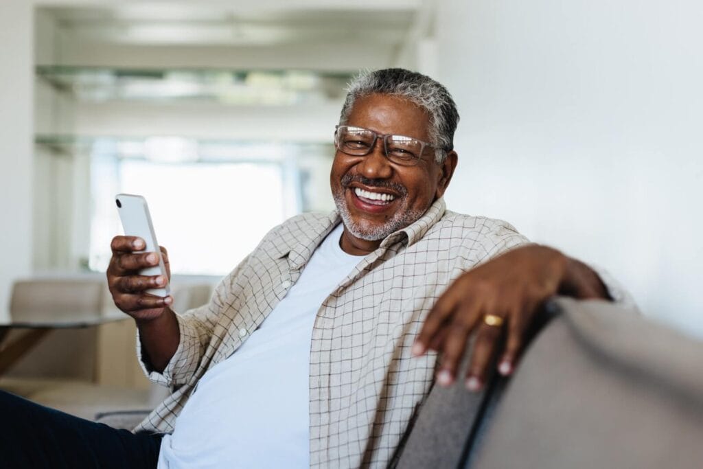 An older adult man is sitting on a couch, smiling at the camera while holding a smartphone.