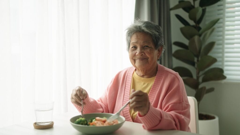 An older adult woman sits at a table. She is holding a spoon and a fork above a bowl of healthy-looking food. There is also a glass of water on the table. She is smiling at the camera.