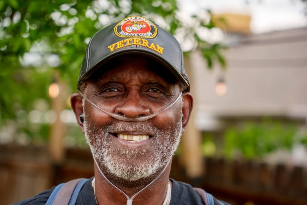 An older adult man is smiling at the camera. He is wearing a nasal cannula for oxygen and a United States Marine Corps baseball hat.