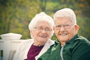 An older adult woman and man sit outside. There are trees behind them. The man is wearing a nasal cannula for oxygen.