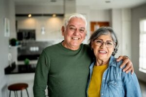 An older adult man and woman are standing in their home. They have their arms around each other's shoulders and smile at the camera.