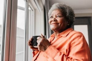 An older adult woman holding a cup of coffee looks out a window in her home, smiling.