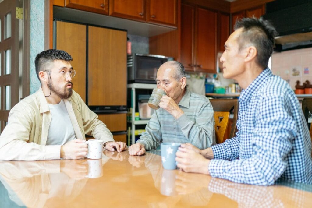 Three men, one of them an older adult, sit at a table drinking coffee and having a discussion.