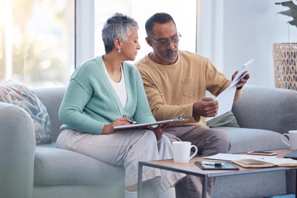 An older adult man and woman sit on a couch, looking over paperwork together.